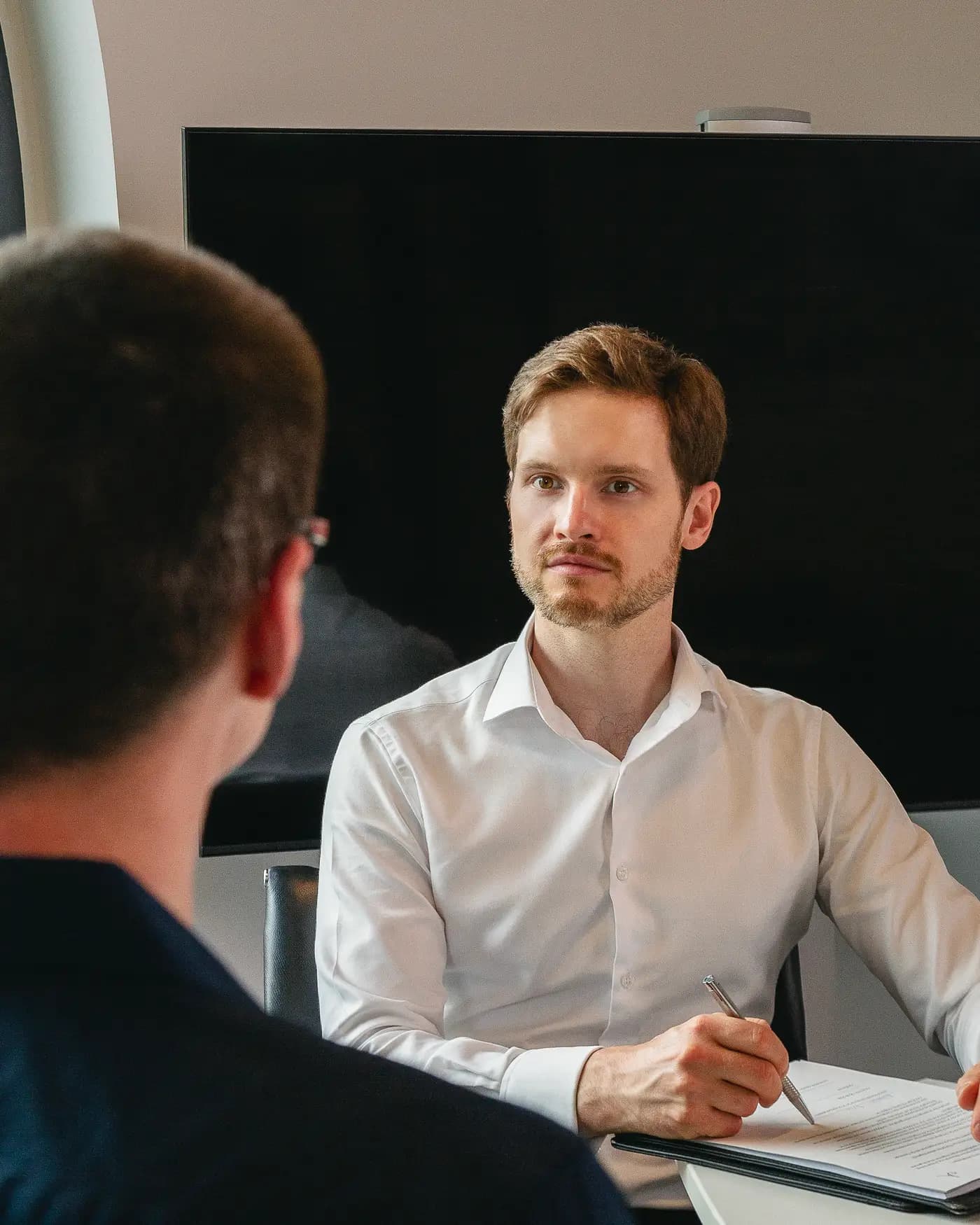 Zwei Personen am runden Tisch, einander gegenüber; der Raum hinter ihnen liegt im Dunkeln.